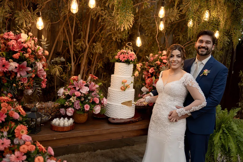 Os noivos diante da parede de flores da recepção, sob os fios de luz.