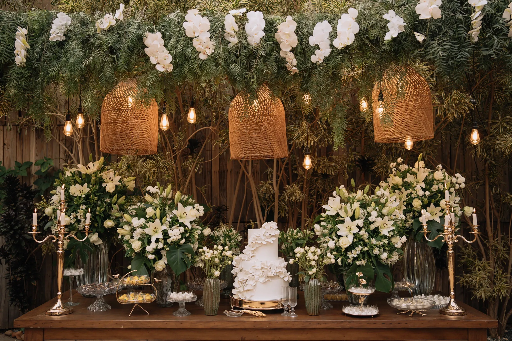 Mesa de bolo sob uma copa de folhagem com orquídeas brancas pendentes, luminárias de palha, castiçais dourados com velas acesas e arranjos altos de lírios.